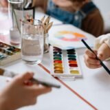 Mâini care pictează cu acuarele pe o foaie albă, pe o masă cu pensule și borcane cu apă