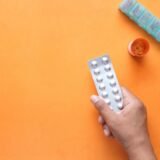 Prospect de medicament desfăcut pe o masă, lângă o folie cu comprimate și o cutie de carton