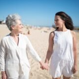 Femeie care ține mâna mamei sale în vârstă, așezate împreună pe o canapea într-o cameră luminoasă